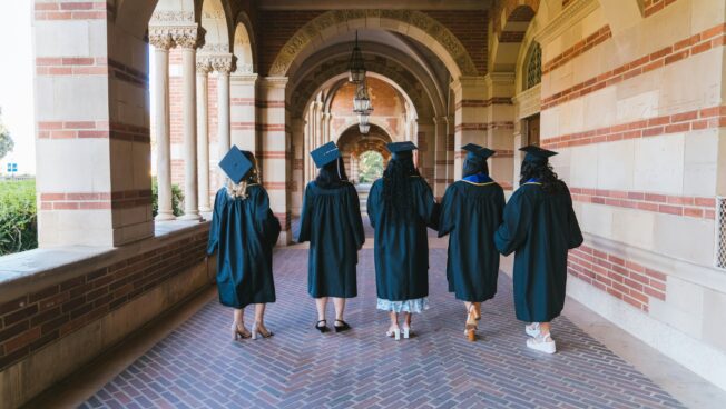 Five young women and graduation gowns and caps walk away from the camera